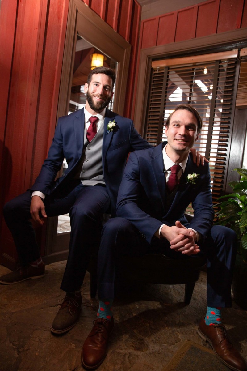Scarlett-Michael-Wedding-Low-Res-51 - TOMMY GATZ ENTERTAINMENT Two men in suits, one seated and one resting his arm on the other's shoulder, pose indoors. They wear formal attire with boutonnieres.