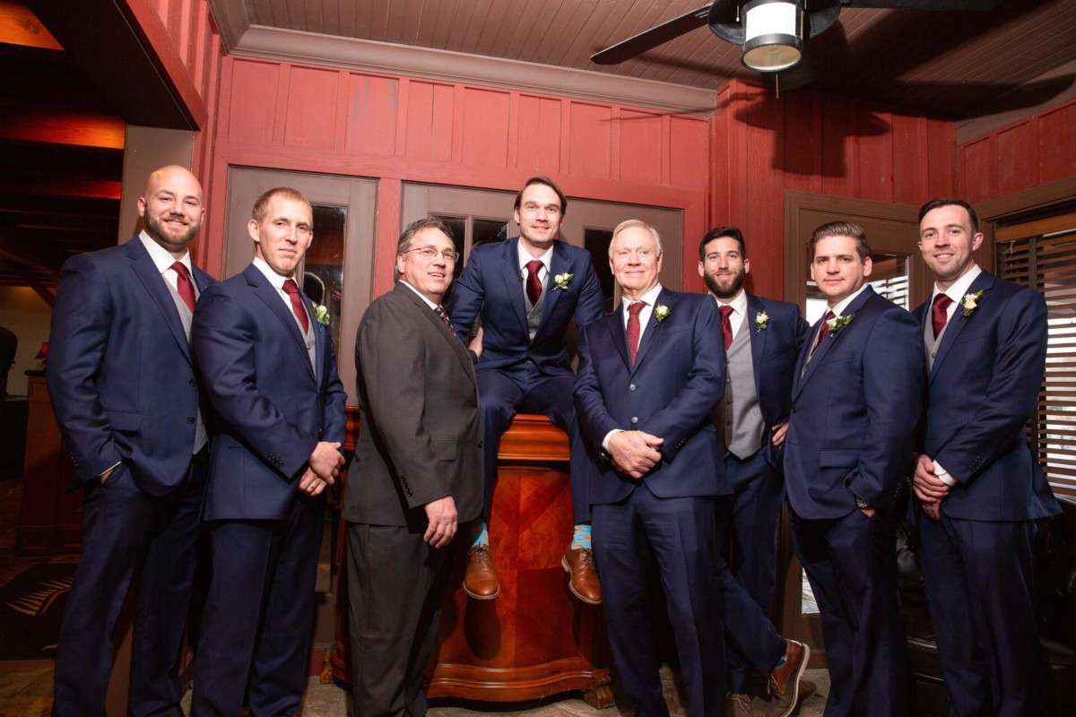 Scarlett-Michael-Wedding-Low-Res-52 - TOMMY GATZ ENTERTAINMENT A group of eight men in formal wear, including blue suits and ties, pose in a room with wood-paneled walls. One man sits on a bar counter while the others stand around him.