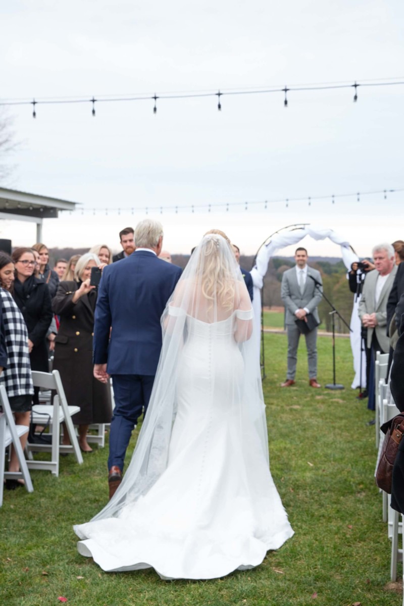 Scarlett-Michael-Wedding-Low-Res-55 - TOMMY GATZ ENTERTAINMENT Bride in a white gown walks with a man in a suit down an outdoor aisle surrounded by seated guests, approaching an archway decorated for a wedding ceremony.