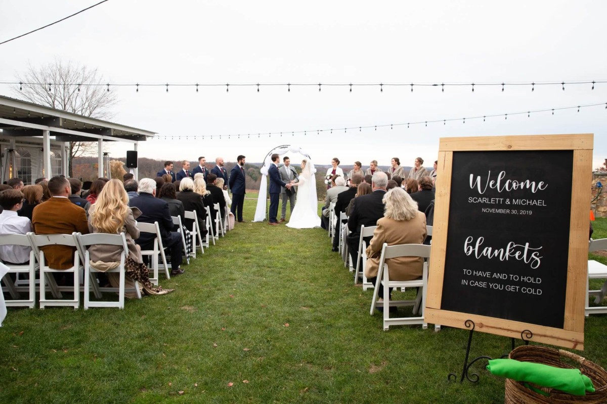 Scarlett-Michael-Wedding-Low-Res-56 - TOMMY GATZ ENTERTAINMENT Outdoor wedding ceremony with guests seated on white chairs, a couple at the altar, and a sign offering blankets.