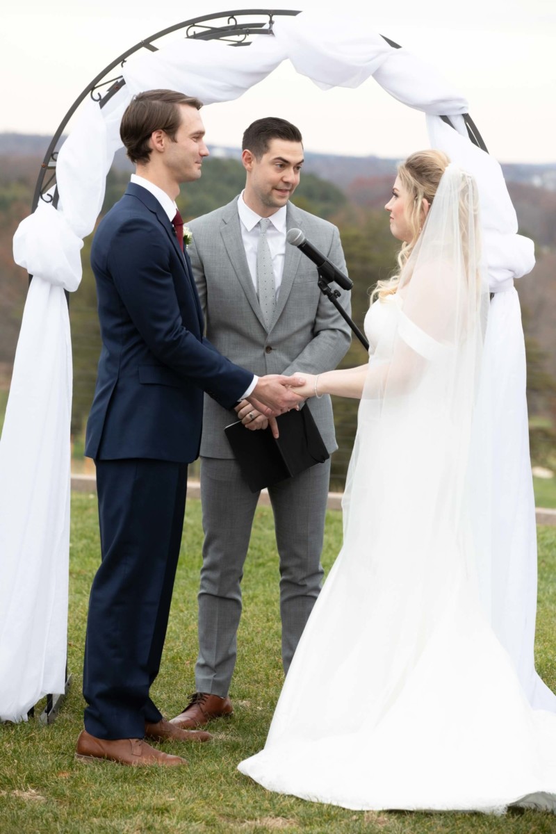 Scarlett-Michael-Wedding-Low-Res-57 - TOMMY GATZ ENTERTAINMENT A bride and groom stand under a white arch holding hands, while a man in a gray suit officiates the ceremony outdoors.