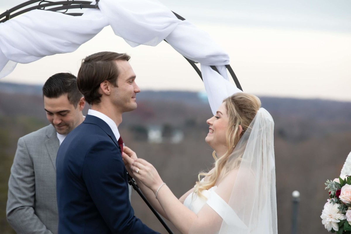 Scarlett-Michael-Wedding-Low-Res-59 - TOMMY GATZ ENTERTAINMENT A couple stands under an arch during their wedding ceremony, with a person in a suit nearby. The bride adjusts the groom's attire.