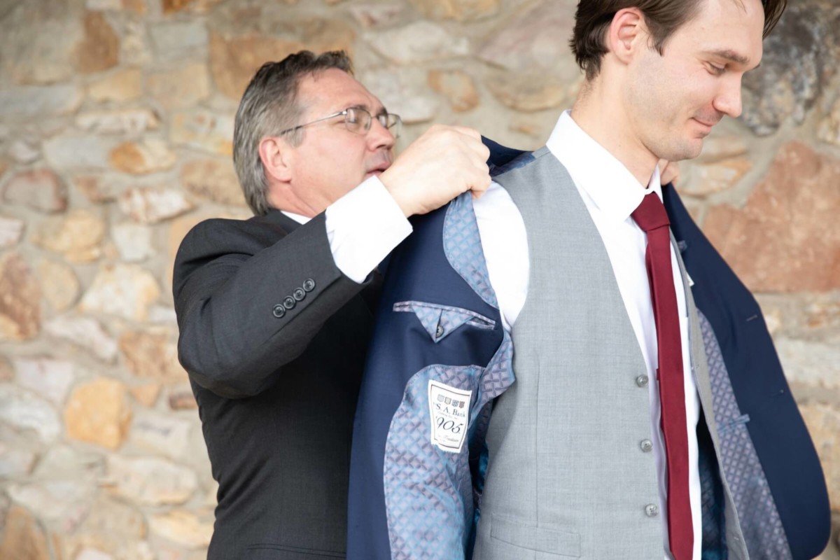 Scarlett-Michael-Wedding-Low-Res-6 - TOMMY GATZ ENTERTAINMENT An older man helps a younger man put on a suit jacket against a stone wall background.