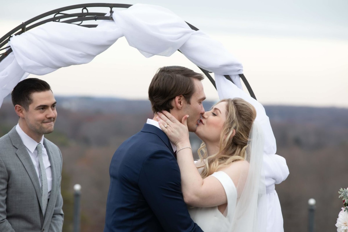 Scarlett-Michael-Wedding-Low-Res-60 - TOMMY GATZ ENTERTAINMENT A couple kisses under a white draped arch during an outdoor wedding ceremony, with a man in a gray suit standing nearby.