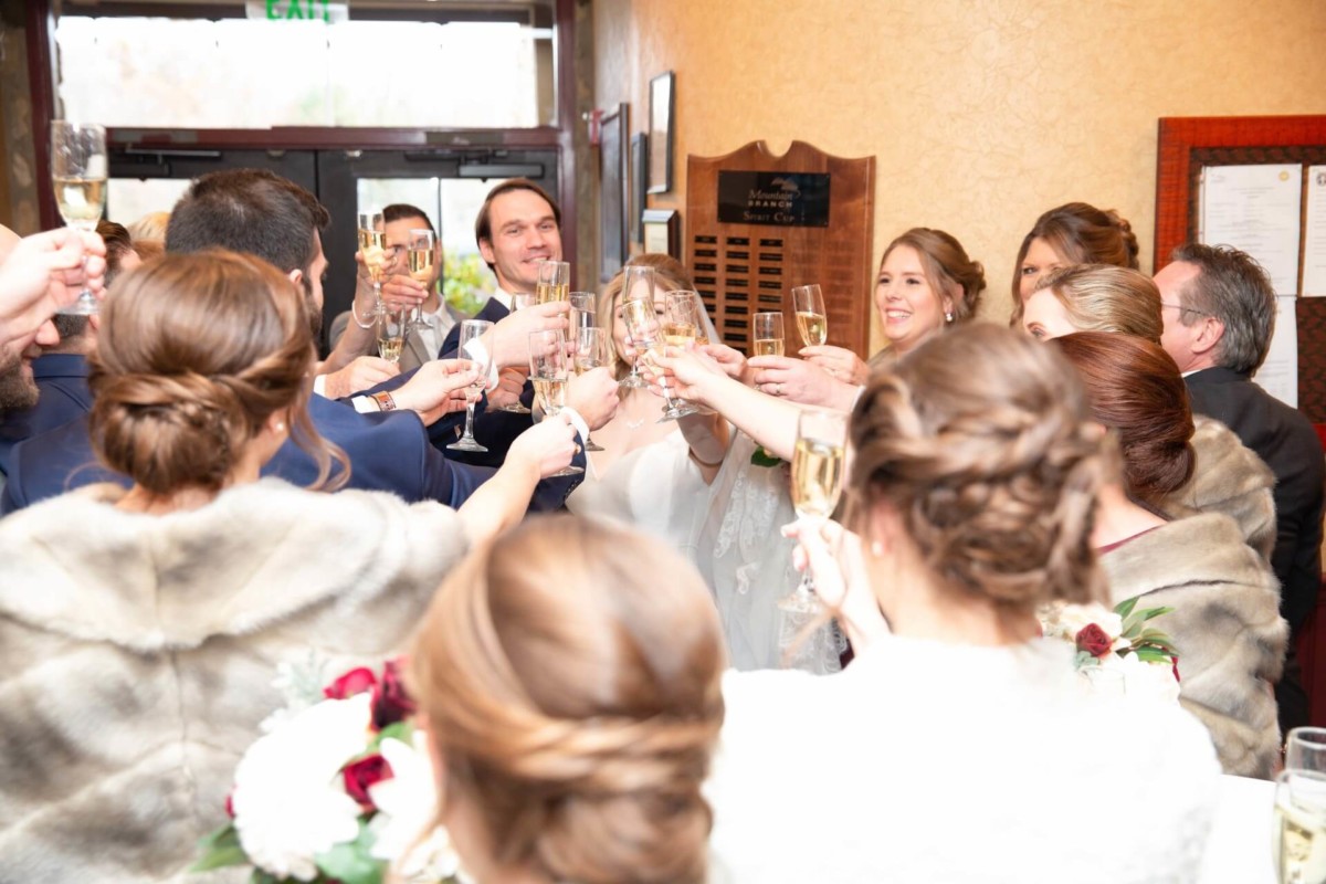 Scarlett-Michael-Wedding-Low-Res-61 - TOMMY GATZ ENTERTAINMENT A group of people raising glasses in a celebratory toast, indoors with a plaque visible on the wall.