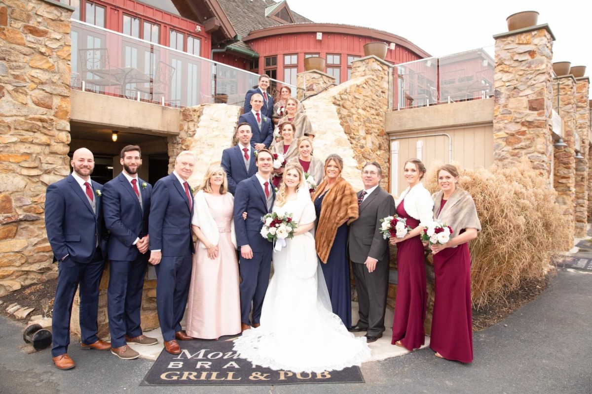Scarlett-Michael-Wedding-Low-Res-63 - TOMMY GATZ ENTERTAINMENT A wedding party poses on stone steps in front of a rustic building. The bride and groom stand in the center, surrounded by their family and friends. Everyone is dressed in formal attire.