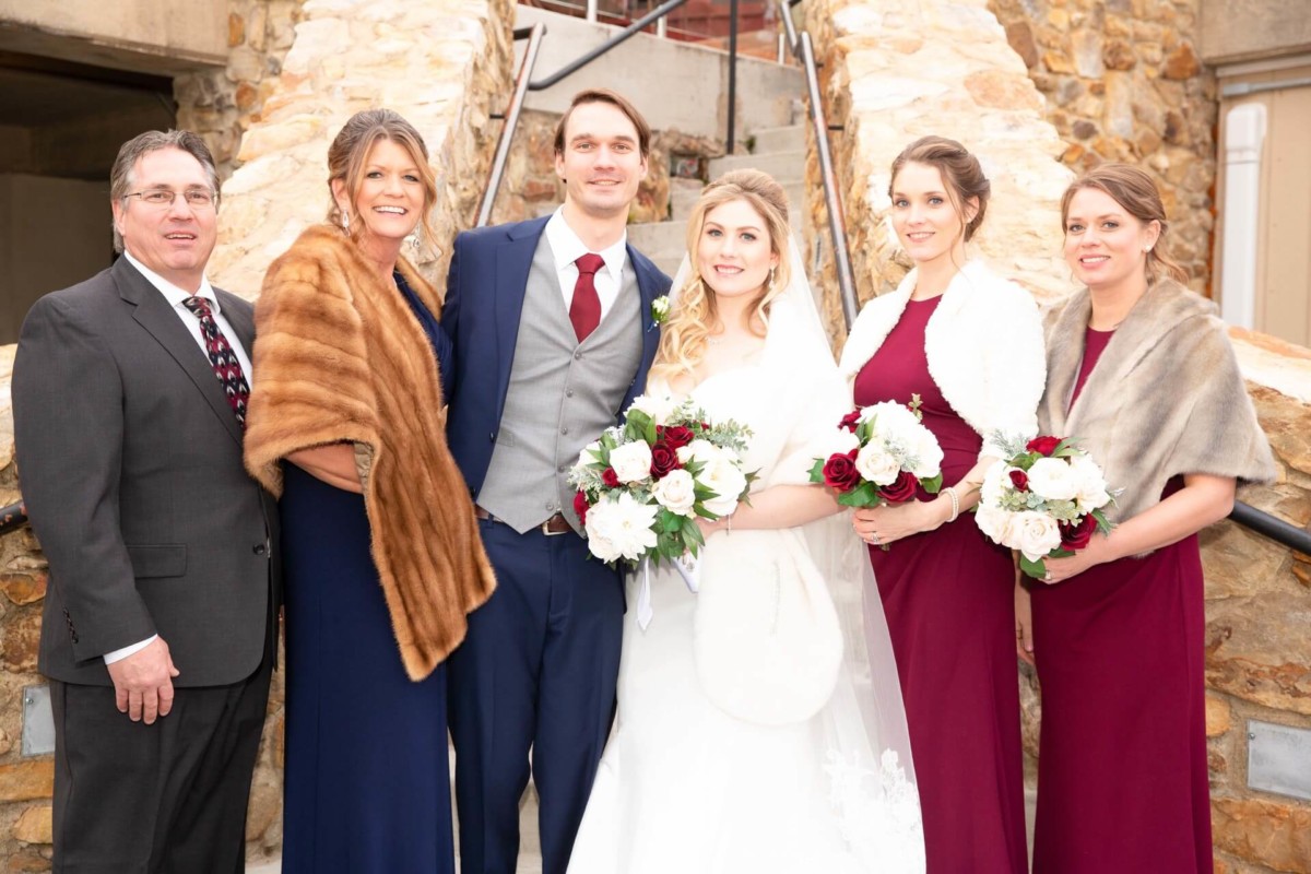 Scarlett-Michael-Wedding-Low-Res-65 - TOMMY GATZ ENTERTAINMENT A group photo of six people dressed in formal attire, including a bride and groom, standing outdoors in front of a stone staircase. Three women are holding bouquets of flowers.