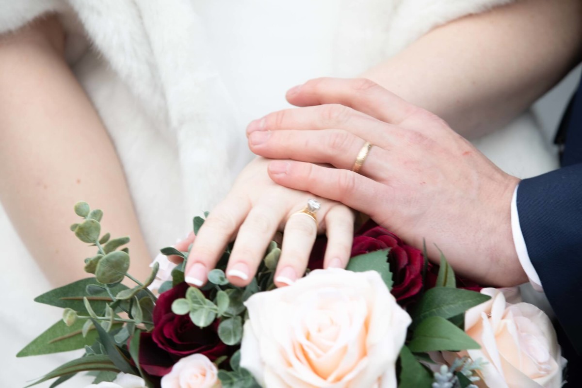 Scarlett-Michael-Wedding-Low-Res-67 - TOMMY GATZ ENTERTAINMENT Close-up of a couple's hands showing wedding rings, resting on a bouquet of pink and maroon flowers with greenery.
