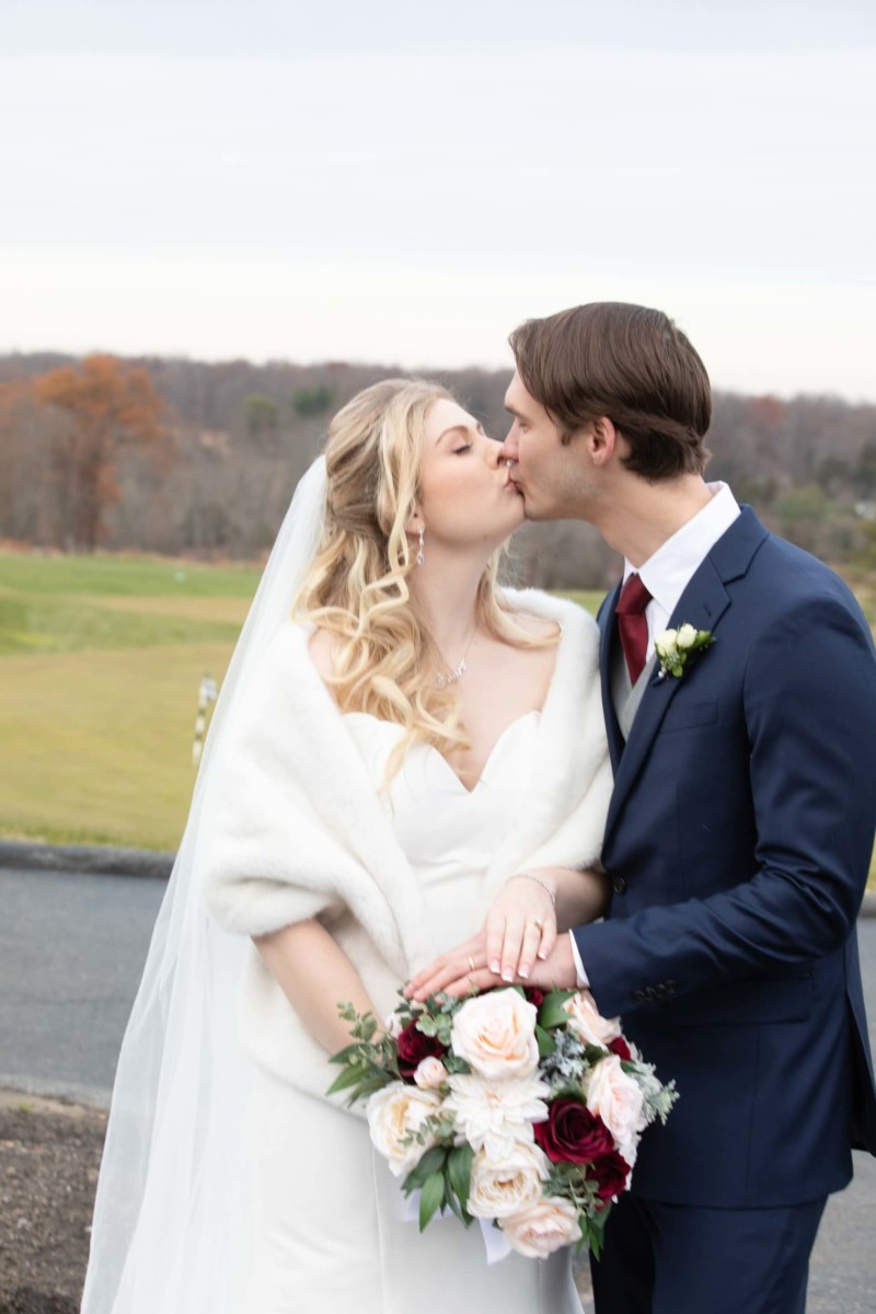 Scarlett-Michael-Wedding-Low-Res-68 - TOMMY GATZ ENTERTAINMENT A couple in wedding attire kisses outdoors, with the bride holding a bouquet of roses.