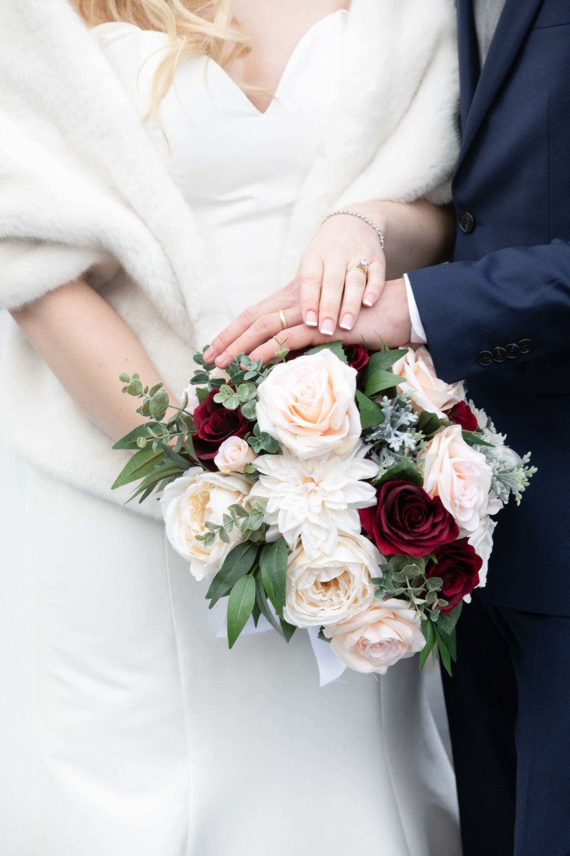 Scarlett-Michael-Wedding-Low-Res-69 - TOMMY GATZ ENTERTAINMENT Bride and groom in formal attire hold hands over a bouquet with white and burgundy roses.