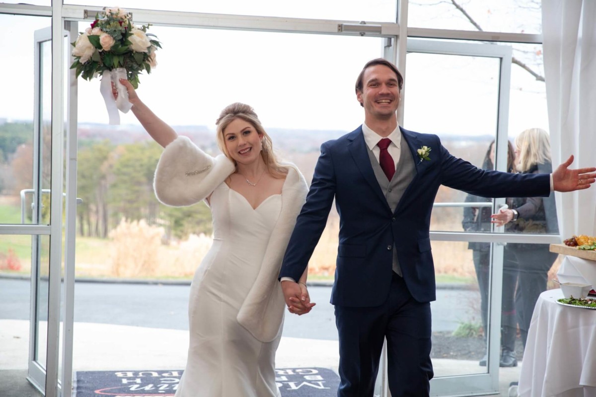 Scarlett-Michael-Wedding-Low-Res-70 - TOMMY GATZ ENTERTAINMENT Bride and groom joyfully entering a venue, holding hands. The bride holds a bouquet in her raised arm, and both are smiling.