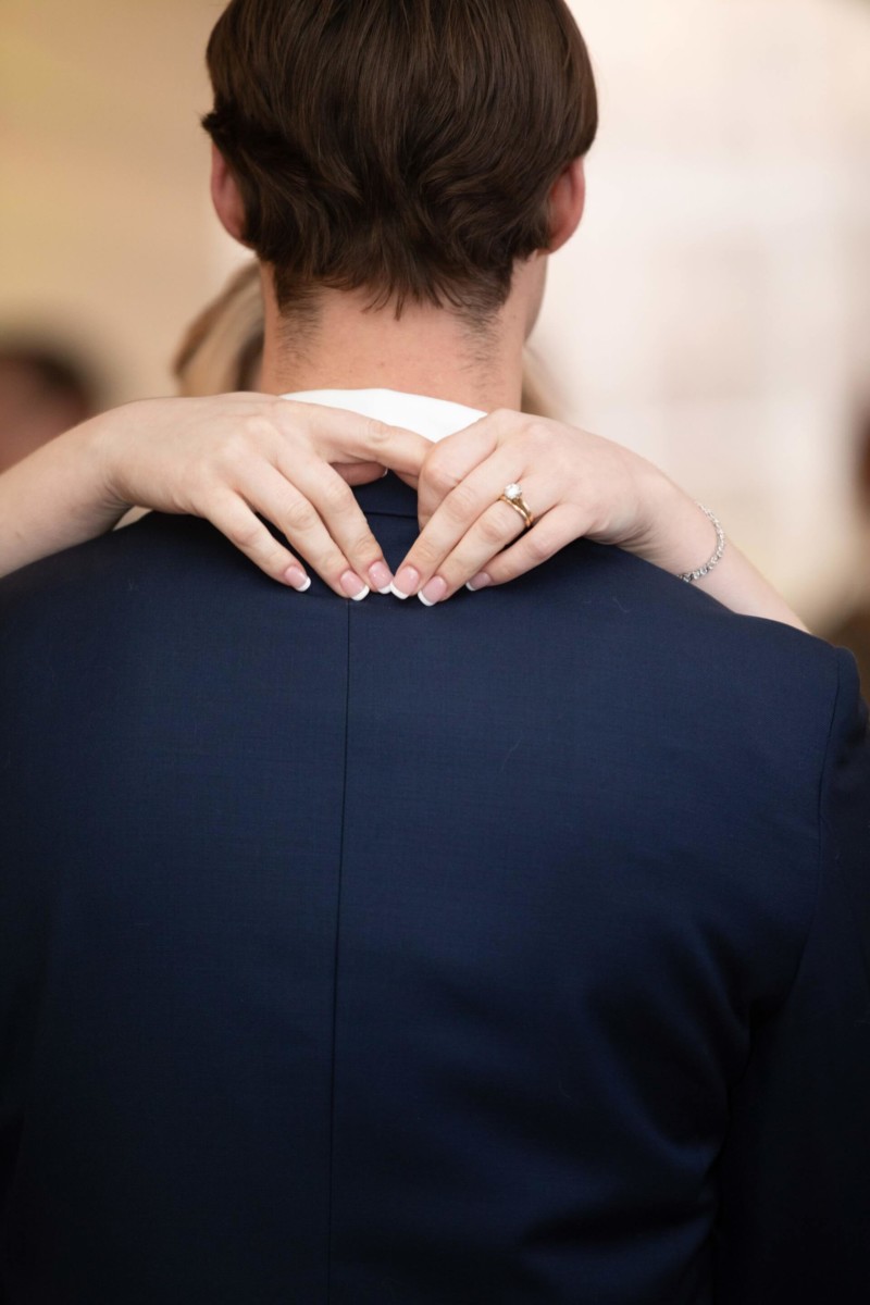 Scarlett-Michael-Wedding-Low-Res-72 - TOMMY GATZ ENTERTAINMENT A person with short hair is seen from behind, wearing a dark suit. Two hands with a ring and bracelet are gently resting on the person's shoulders.
