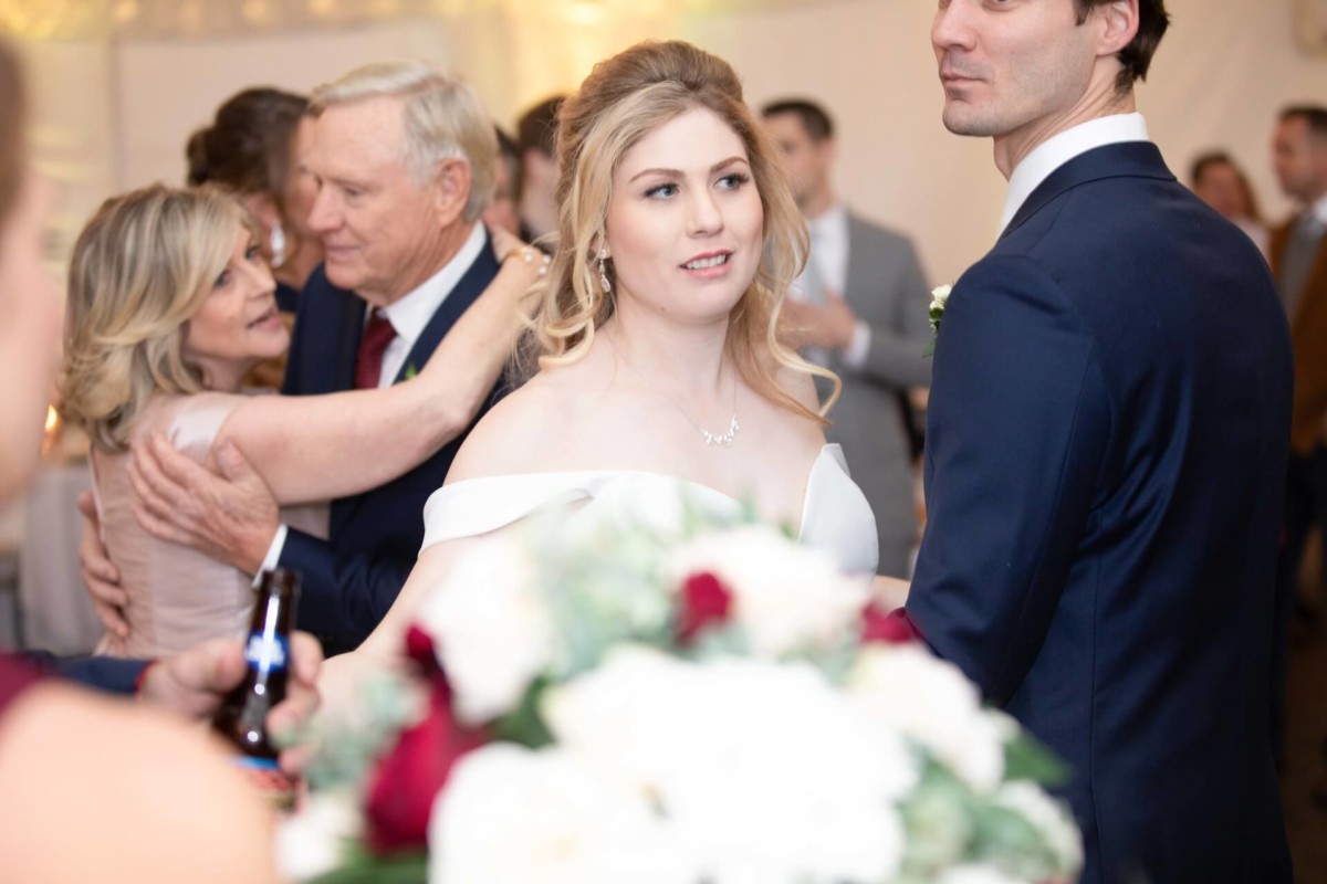 Scarlett-Michael-Wedding-Low-Res-73 - TOMMY GATZ ENTERTAINMENT A bride and groom stand together at a reception. The bride holds a bouquet with red and white flowers. Guests are seen in the background, with some dancing and embracing.