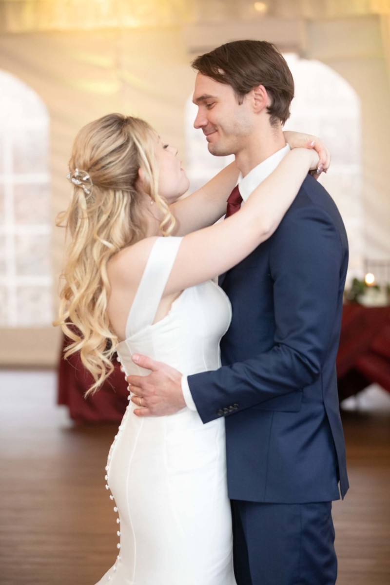 Scarlett-Michael-Wedding-Low-Res-75 - TOMMY GATZ ENTERTAINMENT A bride and groom dance together in a ballroom, with the bride wearing a white gown and the groom in a navy suit.