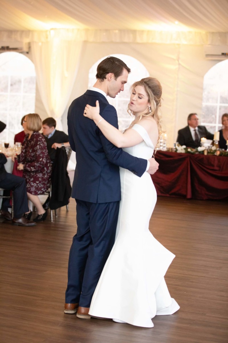 Scarlett-Michael-Wedding-Low-Res-76 - TOMMY GATZ ENTERTAINMENT A couple dances closely in a formal setting. The woman wears a white dress, and the man is in a navy suit. Seated guests are visible in the background within a decorated venue.