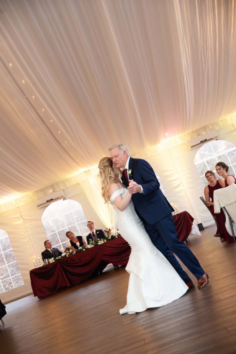 Scarlett-Michael-Wedding-Low-Res-80 - TOMMY GATZ ENTERTAINMENT A bride and an older man dance together inside a tented venue, with seated guests watching in the background.