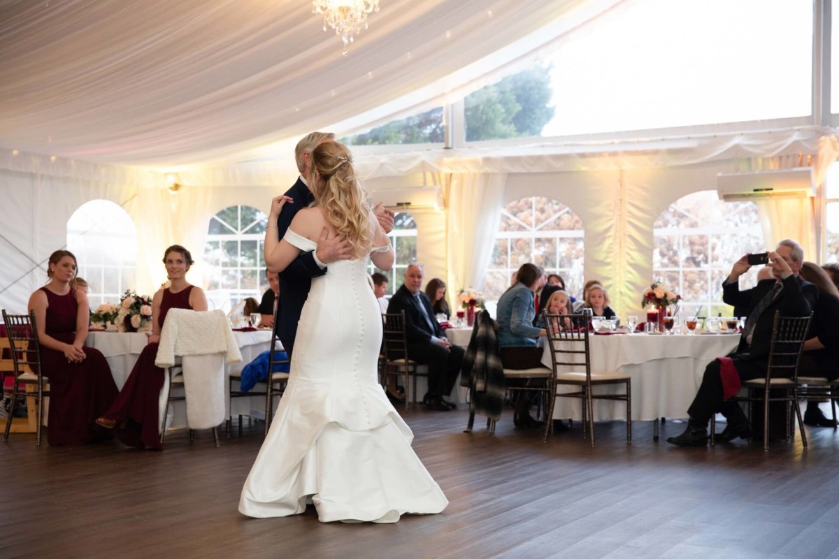 Scarlett-Michael-Wedding-Low-Res-81 - TOMMY GATZ ENTERTAINMENT A couple dances in a white tented venue while guests seated at round tables watch. The bride wears a white gown, and some onlookers are wearing formal attire.
