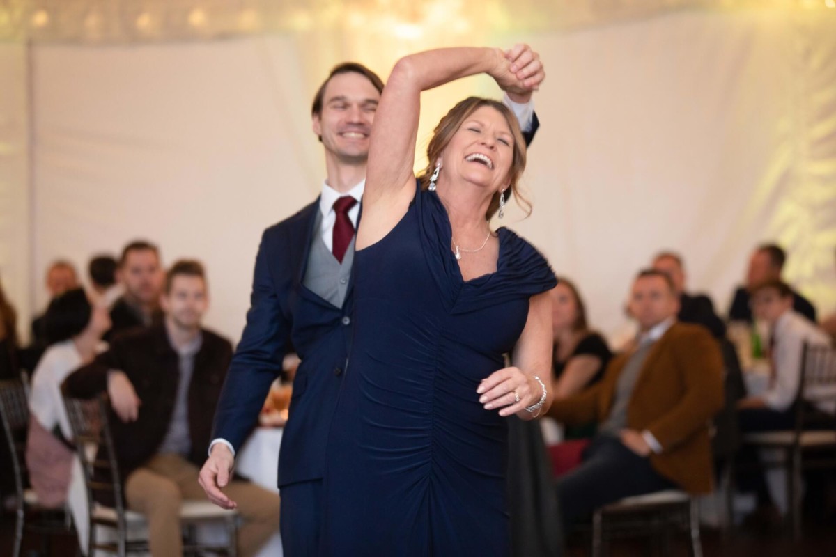 Scarlett-Michael-Wedding-Low-Res-84 - TOMMY GATZ ENTERTAINMENT A woman in a navy dress and a man in a suit dance joyfully in a decorated venue with seated guests in the background.