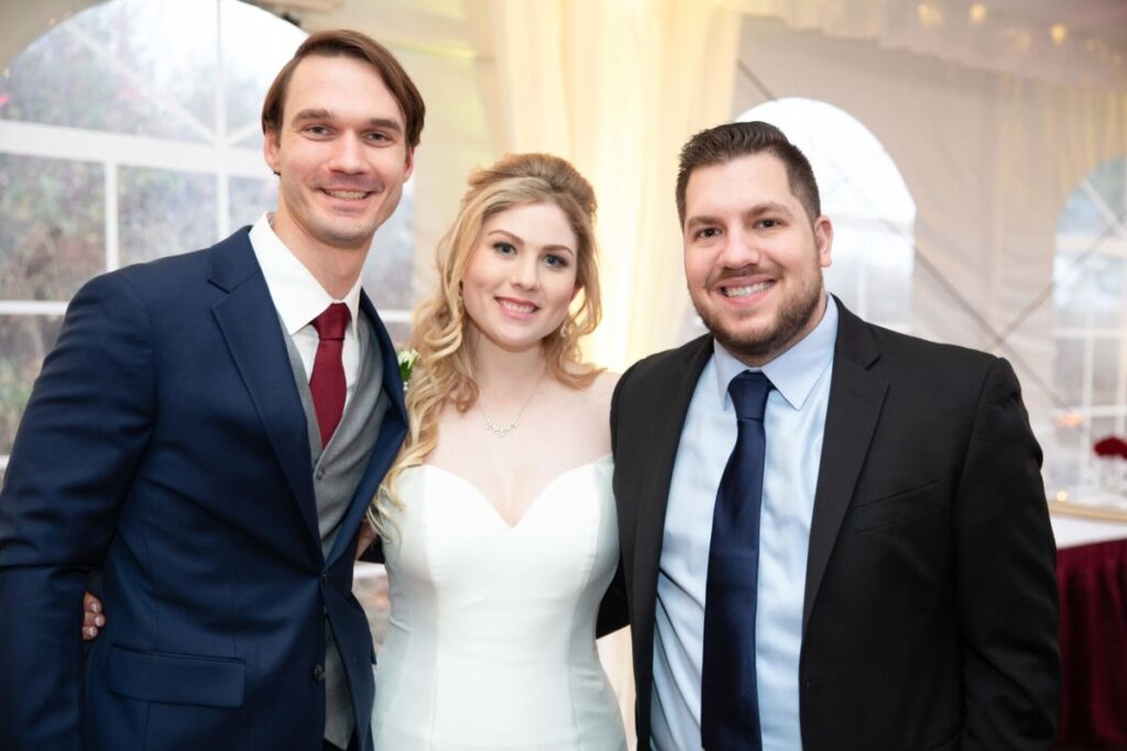 Scarlett-Michael-Wedding-Low-Res-85 - TOMMY GATZ ENTERTAINMENT Three people dressed formally smile at the camera; a woman in a wedding dress is flanked by two men in suits, standing inside a decorated venue.