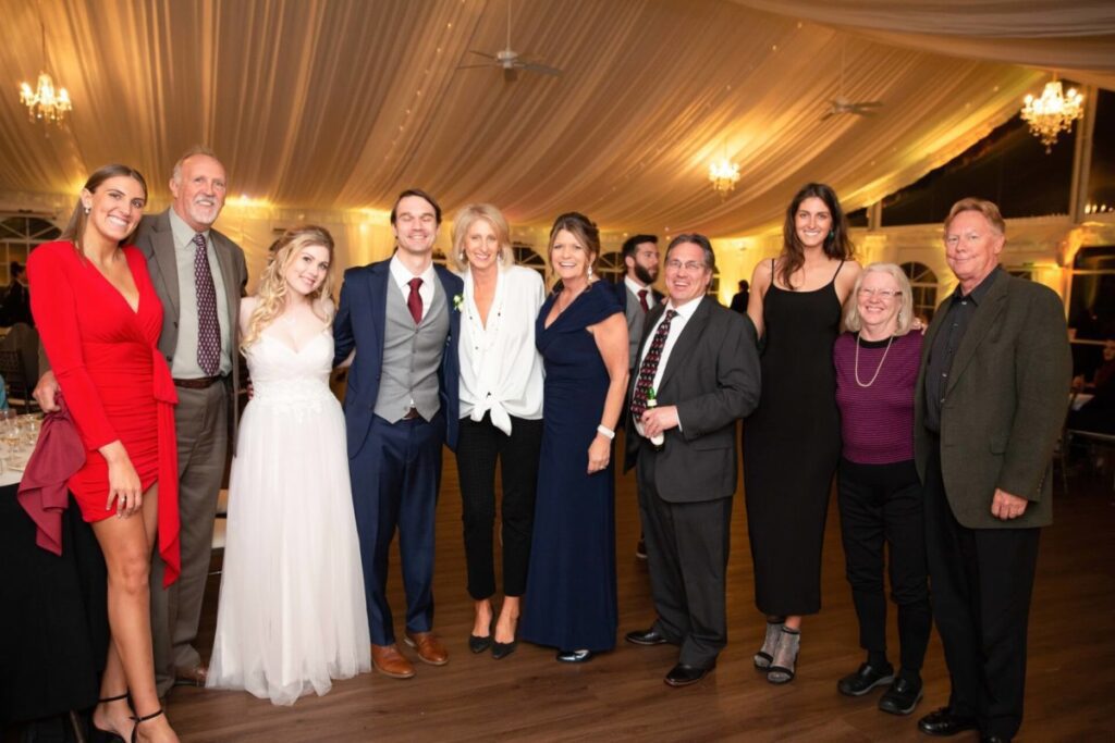 Scarlett-Michael-Wedding-Low-Res-86 - TOMMY GATZ ENTERTAINMENT A group of people in formal attire posing together at an indoor event with chandeliers and draped ceilings.