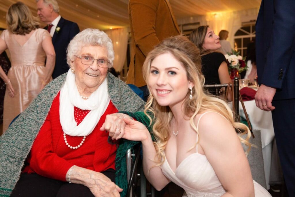 Scarlett-Michael-Wedding-Low-Res-87 - TOMMY GATZ ENTERTAINMENT An elderly woman in a red sweater sits in a wheelchair, smiling, with a young woman in a white dress kneeling beside her, holding her hand. They are indoors at an event.