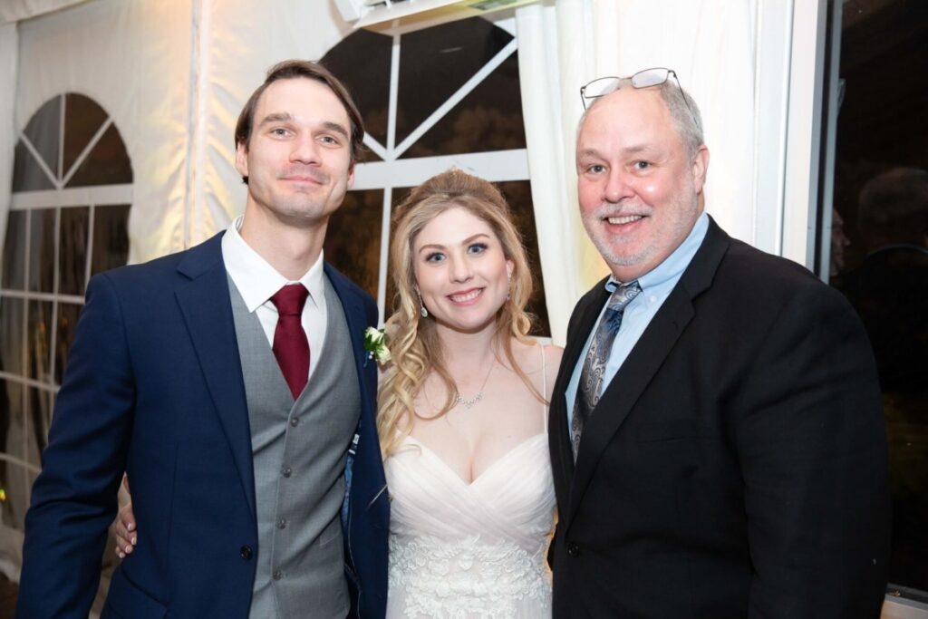 Scarlett-Michael-Wedding-Low-Res-88 - TOMMY GATZ ENTERTAINMENT Three people stand together indoors, dressed in formal attire. The man on the left wears a suit, the woman in a wedding dress, and the man on the right in a suit.