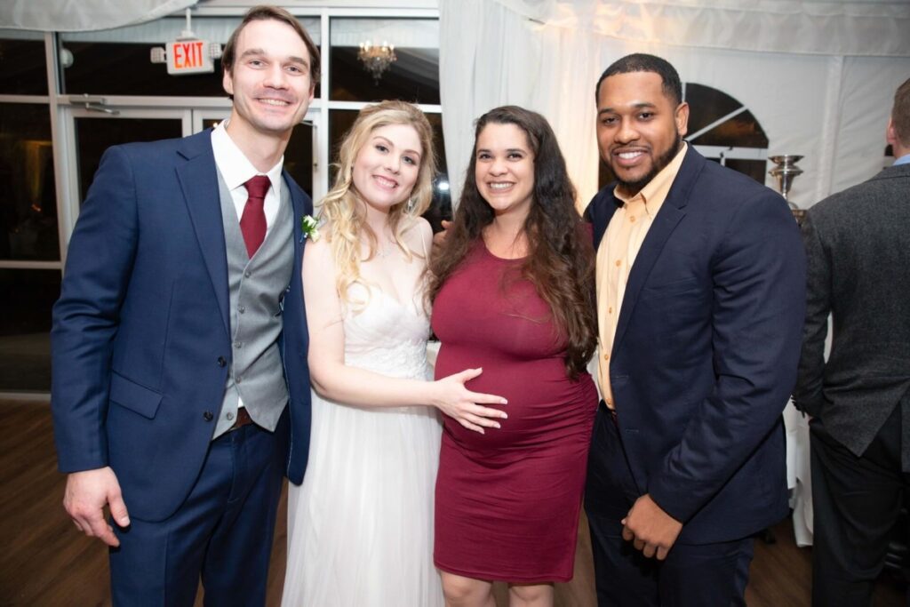 Scarlett-Michael-Wedding-Low-Res-89 - TOMMY GATZ ENTERTAINMENT Four people posing in formal attire at an indoor event. A bride in a white dress and three individuals in suits and a red dress stand smiling.