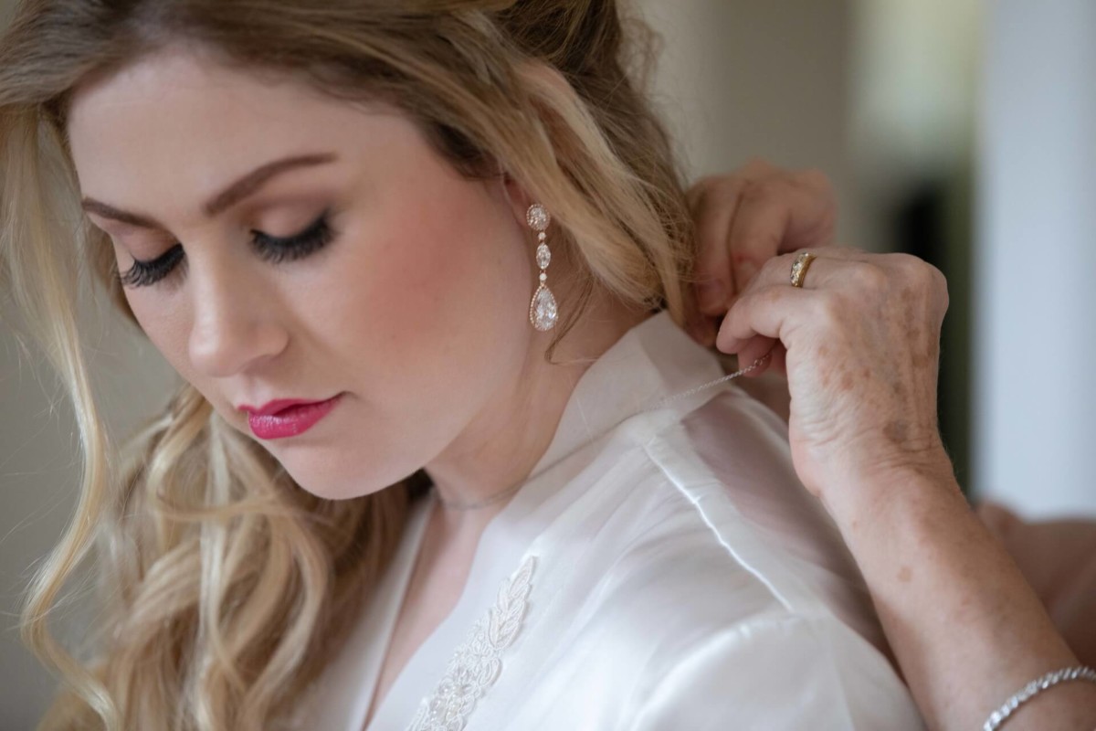 Scarlett-Michael-Wedding-Low-Res-9 - TOMMY GATZ ENTERTAINMENT A bride in a white robe is having her necklace fastened by another person's hand. She has blonde hair and is wearing drop earrings.