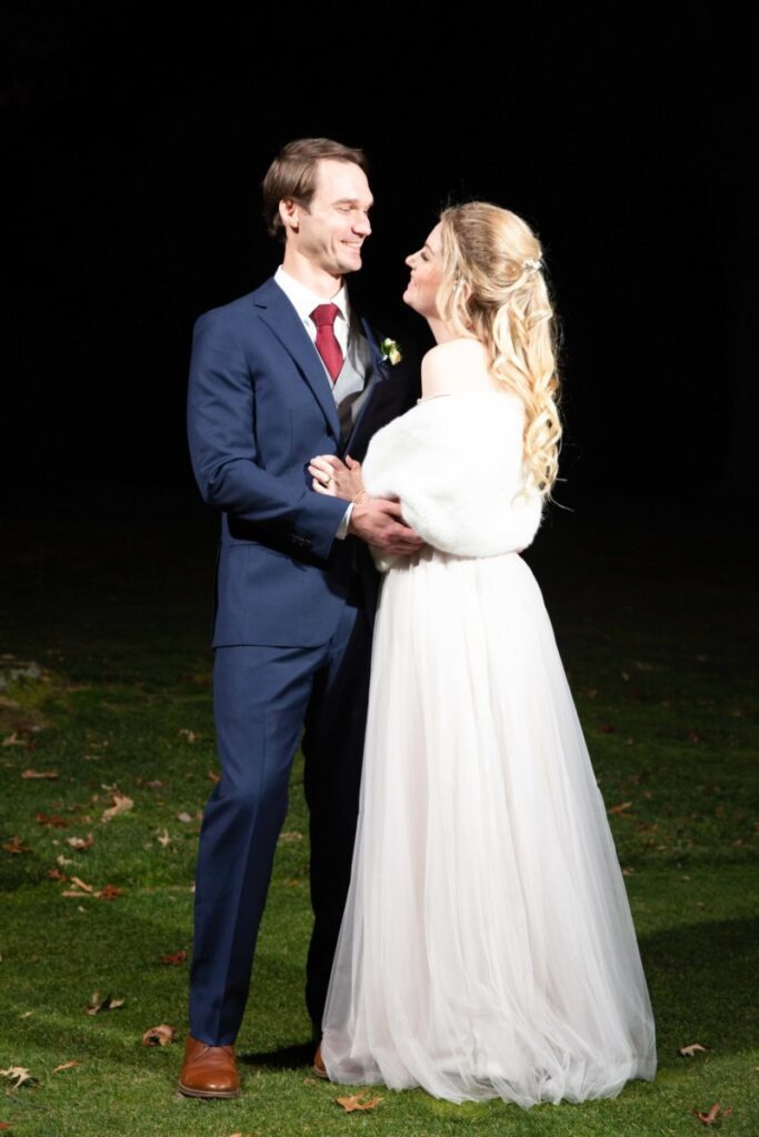 Scarlett-Michael-Wedding-Low-Res-90 - TOMMY GATZ ENTERTAINMENT A couple in formal attire stands on grass at night; the man wears a blue suit and red tie, the woman in a white dress with fur shrug, both looking at each other.