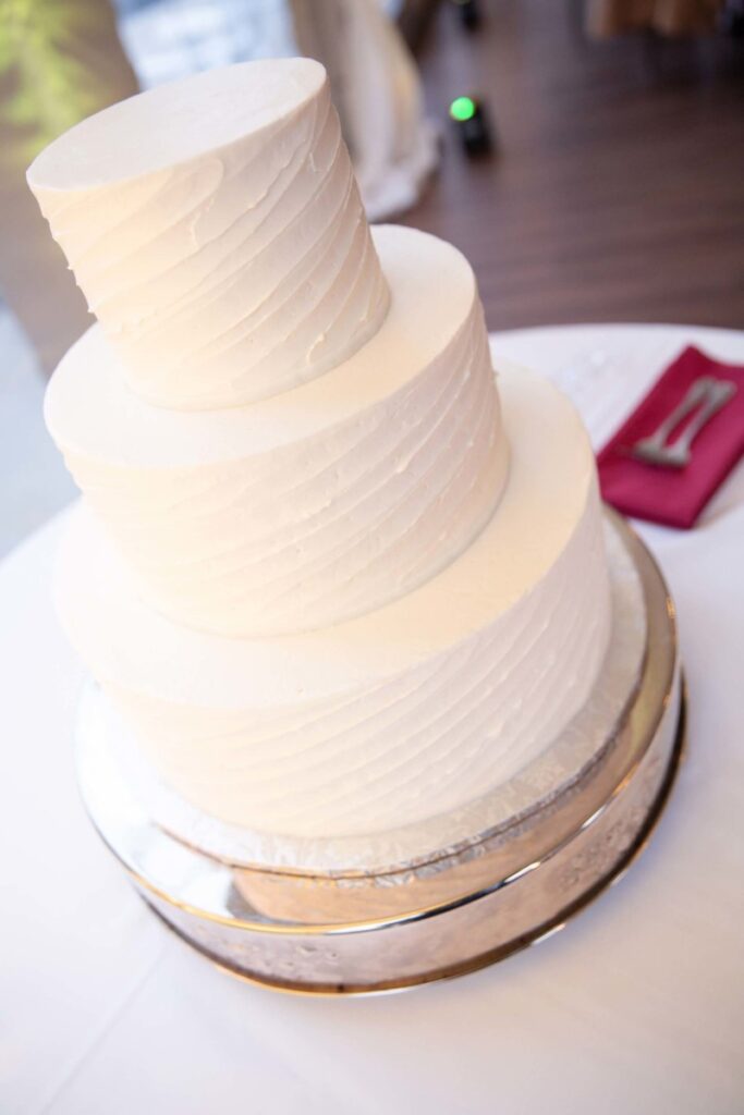 Scarlett-Michael-Wedding-Low-Res-94 - TOMMY GATZ ENTERTAINMENT Three-tiered white frosted cake on a silver stand, placed on a table with a pink napkin and silver fork beside it.