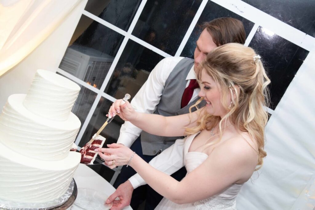 Scarlett-Michael-Wedding-Low-Res-95 - TOMMY GATZ ENTERTAINMENT A bride and groom cut a slice of a three-tiered white wedding cake together.