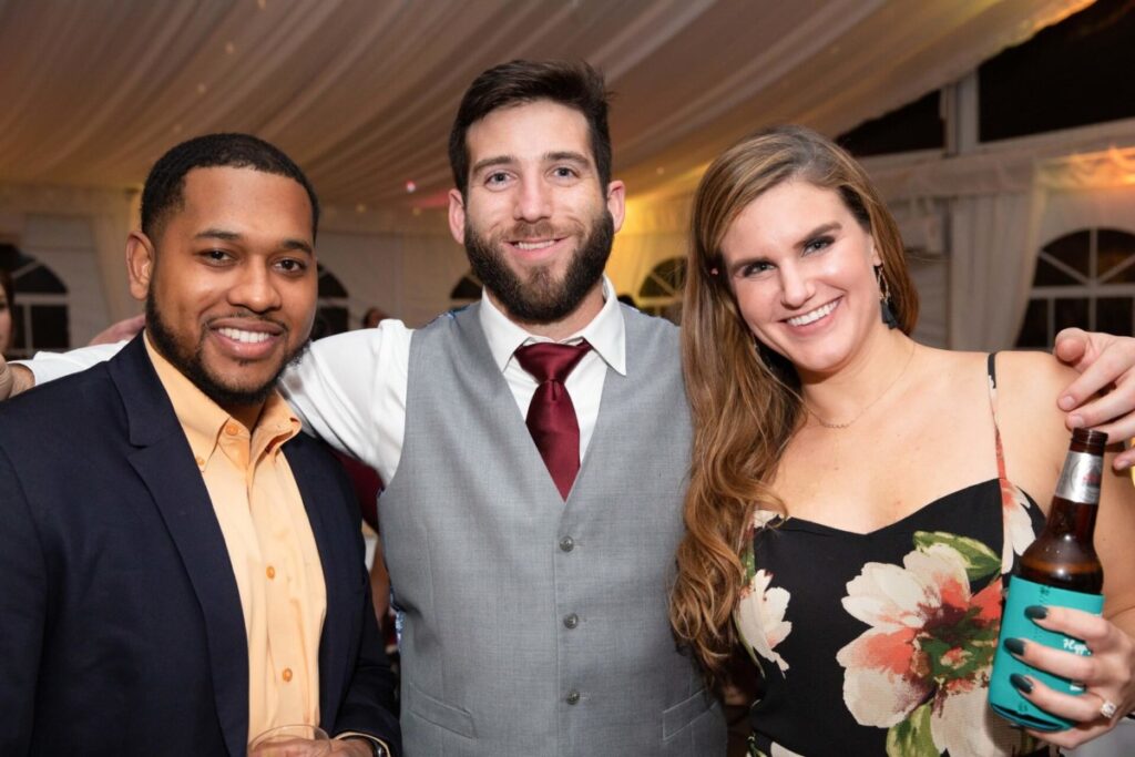 Scarlett-Michael-Wedding-Low-Res-97 - TOMMY GATZ ENTERTAINMENT Three people standing together at an event, smiling at the camera. One holds a drink bottle, and another holds a glass. They're dressed in semi-formal attire.