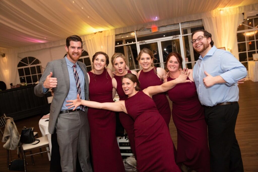 A group of seven people, five women in matching burgundy dresses and two men in suits, pose together indoors, smiling and making playful gestures.
