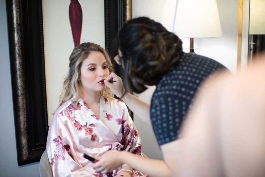 Shannon-James-Wedding-Low-Res-1 - TOMMY GATZ ENTERTAINMENT A woman in a floral robe sits while another person applies makeup to her face. A lamp and framed artwork are in the background.