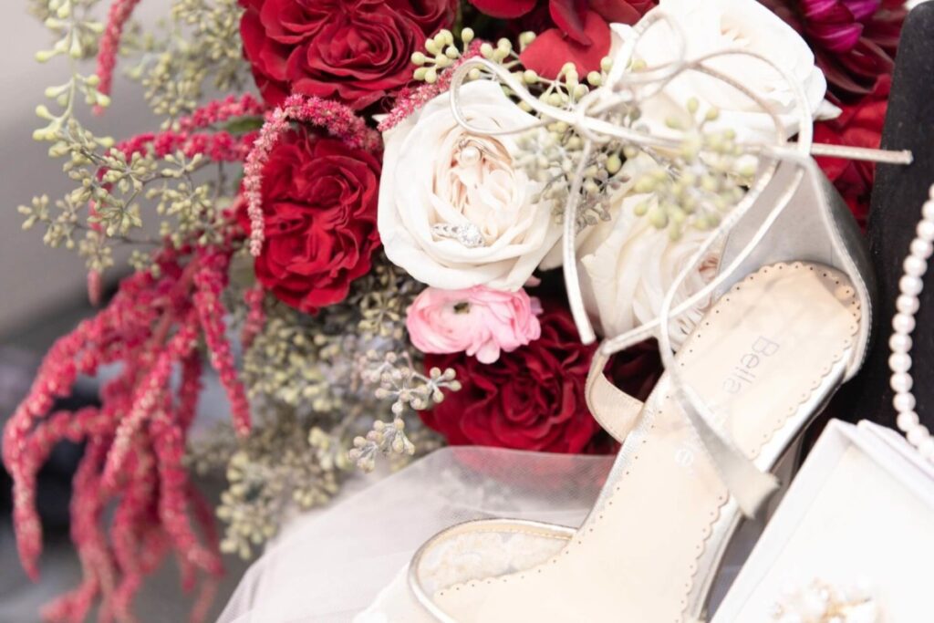 Shannon-James-Wedding-Low-Res-10 - TOMMY GATZ ENTERTAINMENT Close-up of a bridal bouquet with red and white flowers next to a beige high-heeled shoe.