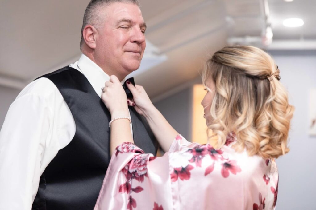 Shannon-James-Wedding-Low-Res-11 - TOMMY GATZ ENTERTAINMENT A woman adjusts a man's bow tie. The man wears a white shirt with a black vest, and the woman is in a floral satin robe. They are indoors with ceiling lights in the background.