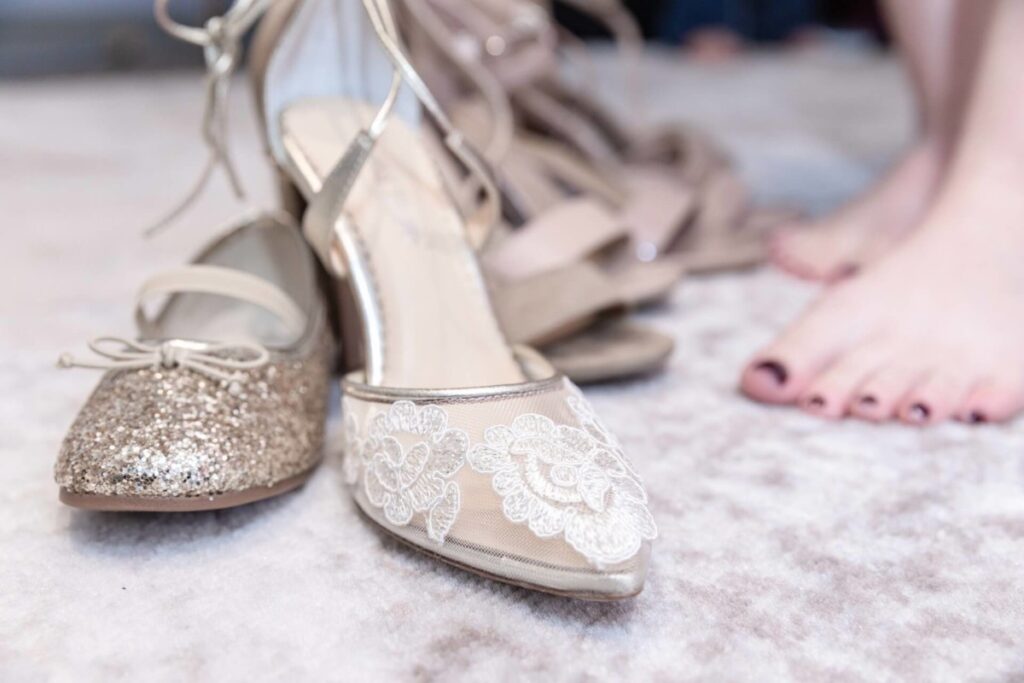 Shannon-James-Wedding-Low-Res-12 - TOMMY GATZ ENTERTAINMENT A selection of women's shoes, including a glittery flat and a lace-trimmed sandal, is displayed on a light carpet next to bare feet.