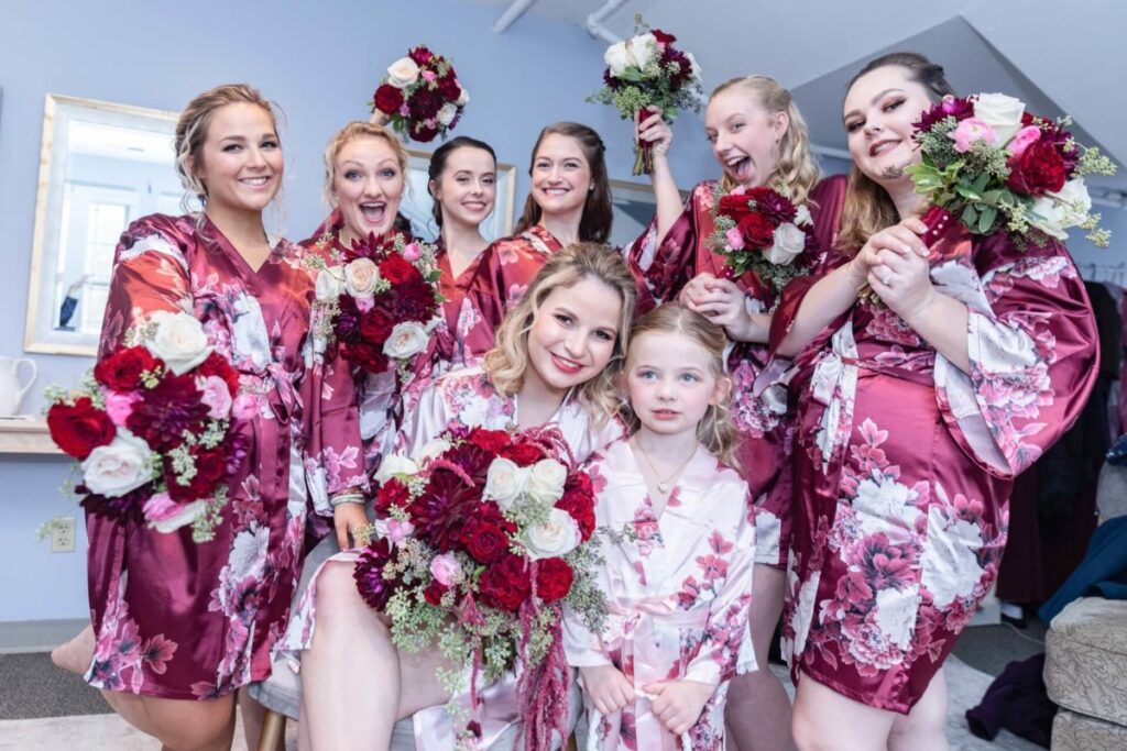Shannon-James-Wedding-Low-Res-13 - TOMMY GATZ ENTERTAINMENT Eight women and one girl in matching floral robes hold bouquets, smiling and posing in a room with light walls and a mirror.