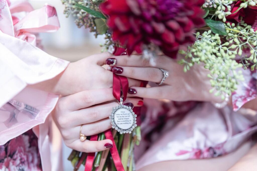 Shannon-James-Wedding-Low-Res-15 - TOMMY GATZ ENTERTAINMENT Close-up of a person holding a bouquet with dark red flowers and a silver pendant. The person has red and white painted nails and is wearing a ring.
