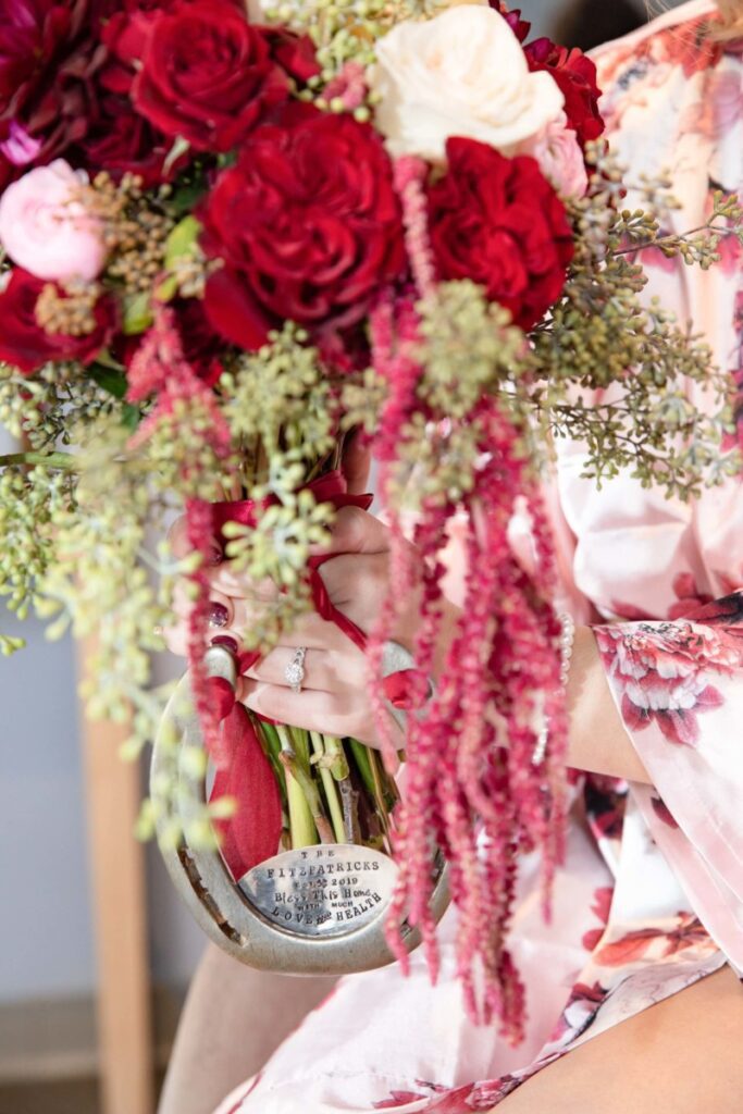 Shannon-James-Wedding-Low-Res-17 - TOMMY GATZ ENTERTAINMENT A person in a floral robe holds a bouquet with red, white, and pink flowers, greenery, and cascading amaranthus, in a clear vase with a nameplate. A ring is visible on their finger.