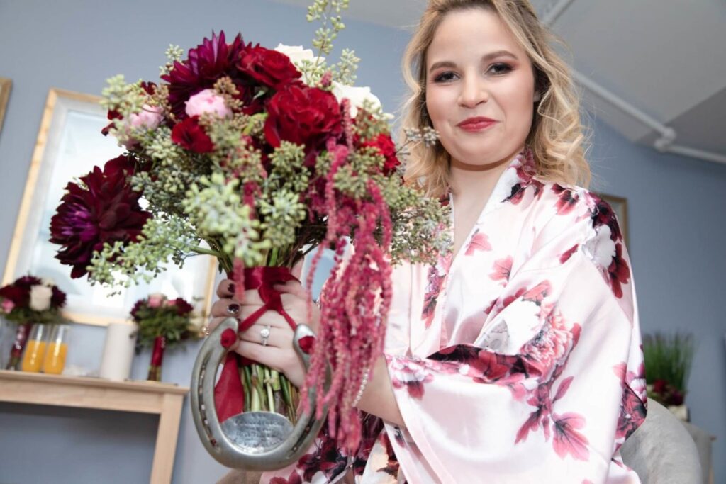 Shannon-James-Wedding-Low-Res-19 - TOMMY GATZ ENTERTAINMENT Person in a floral robe holding a bouquet with red and pink flowers, smiling indoors.