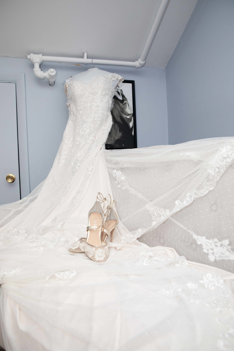 Shannon-James-Wedding-Low-Res-20 - TOMMY GATZ ENTERTAINMENT A lacy white wedding dress is displayed on a blue couch with beige high heels placed in front. A monochrome portrait hangs on the wall.