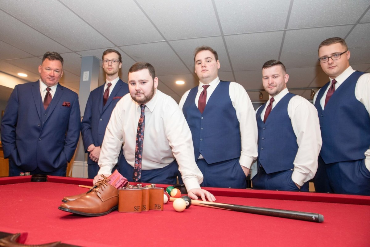 Shannon-James-Wedding-Low-Res-22 - TOMMY GATZ ENTERTAINMENT Six men in suits pose around a red pool table with shoes and a tie displayed on it.