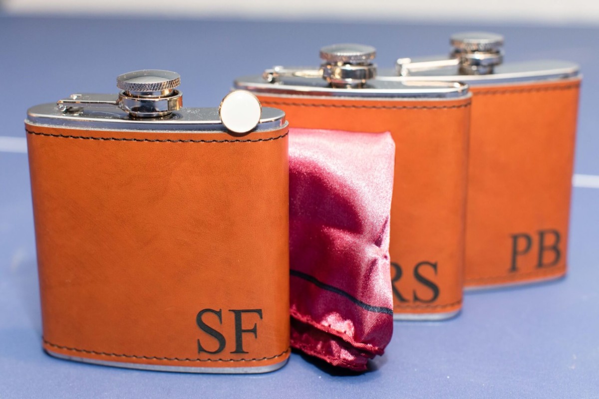 Shannon-James-Wedding-Low-Res-23 - TOMMY GATZ ENTERTAINMENT Three brown leather-wrapped flasks labeled "SF," "RS," and "PB," with a red satin cloth partially covering the middle flask, placed on a blue surface.
