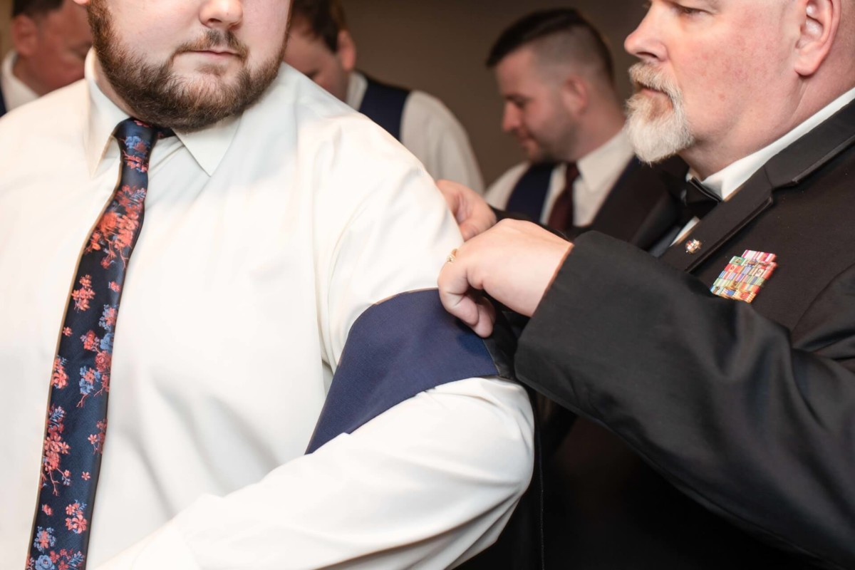 Shannon-James-Wedding-Low-Res-24 - TOMMY GATZ ENTERTAINMENT A man helps another man put on a blue sash. Both are wearing formal attire, and there are other men in the background, also in formal clothing.