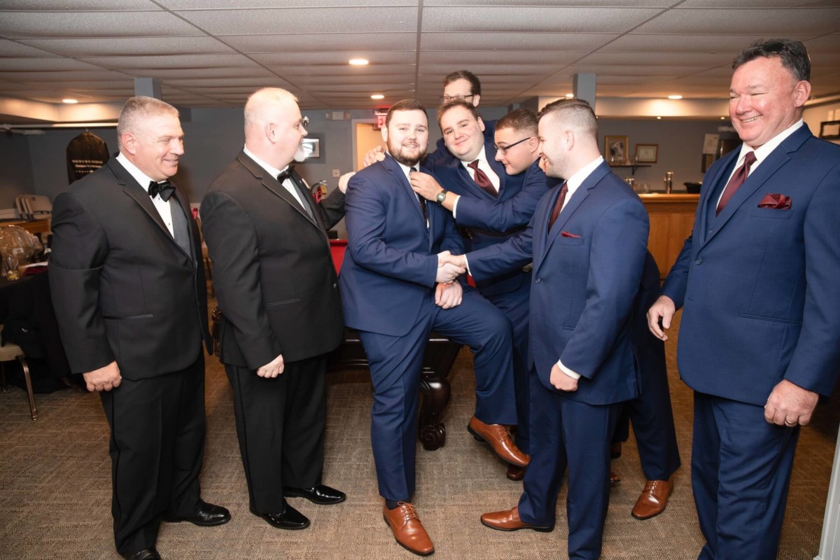 Shannon-James-Wedding-Low-Res-26 - TOMMY GATZ ENTERTAINMENT A group of men in formal suits gather around one man, smiling and adjusting his attire in an indoor setting.