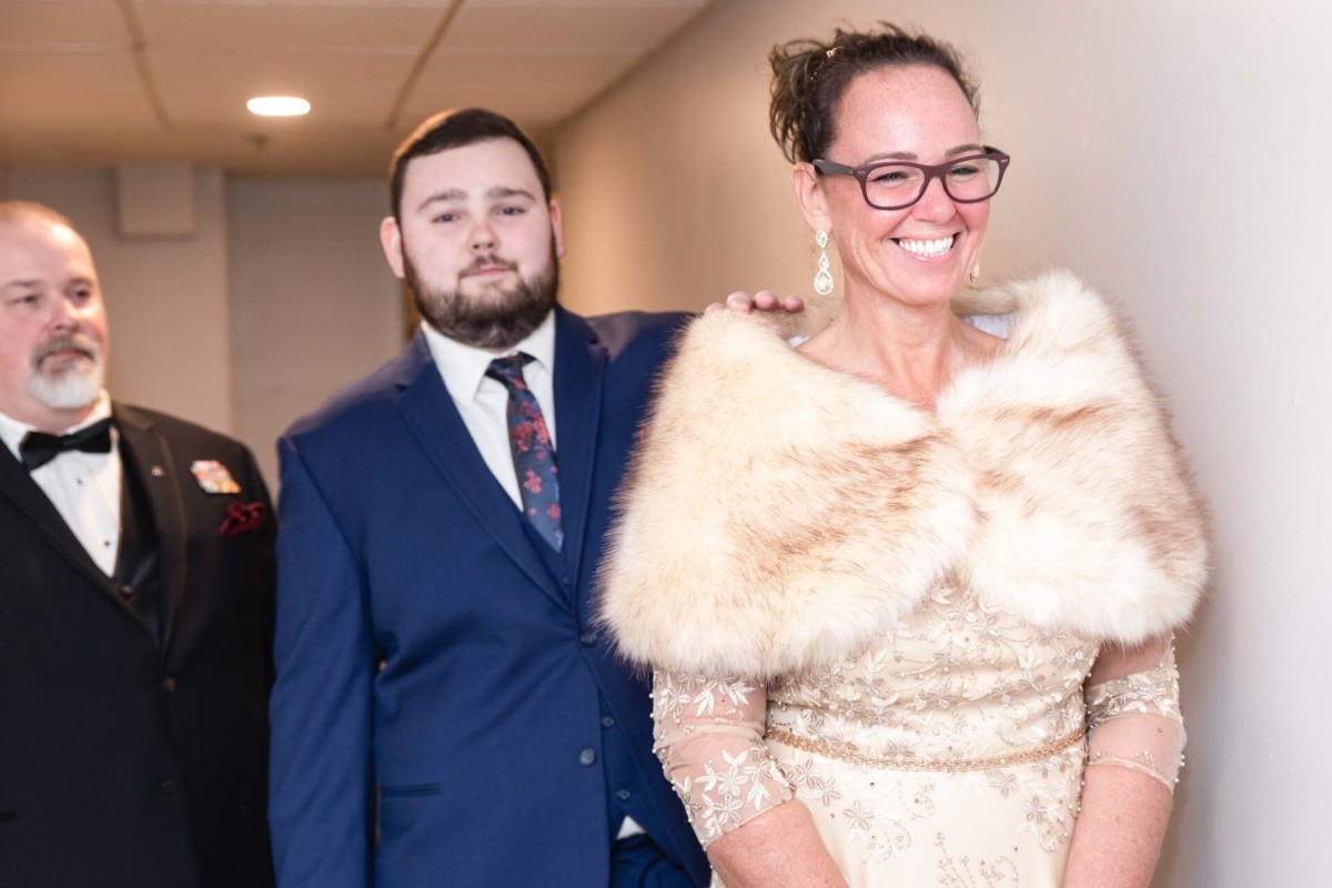 Shannon-James-Wedding-Low-Res-27 - TOMMY GATZ ENTERTAINMENT Three people in formal attire, with a woman wearing a fur stole and glasses smiling in the foreground.