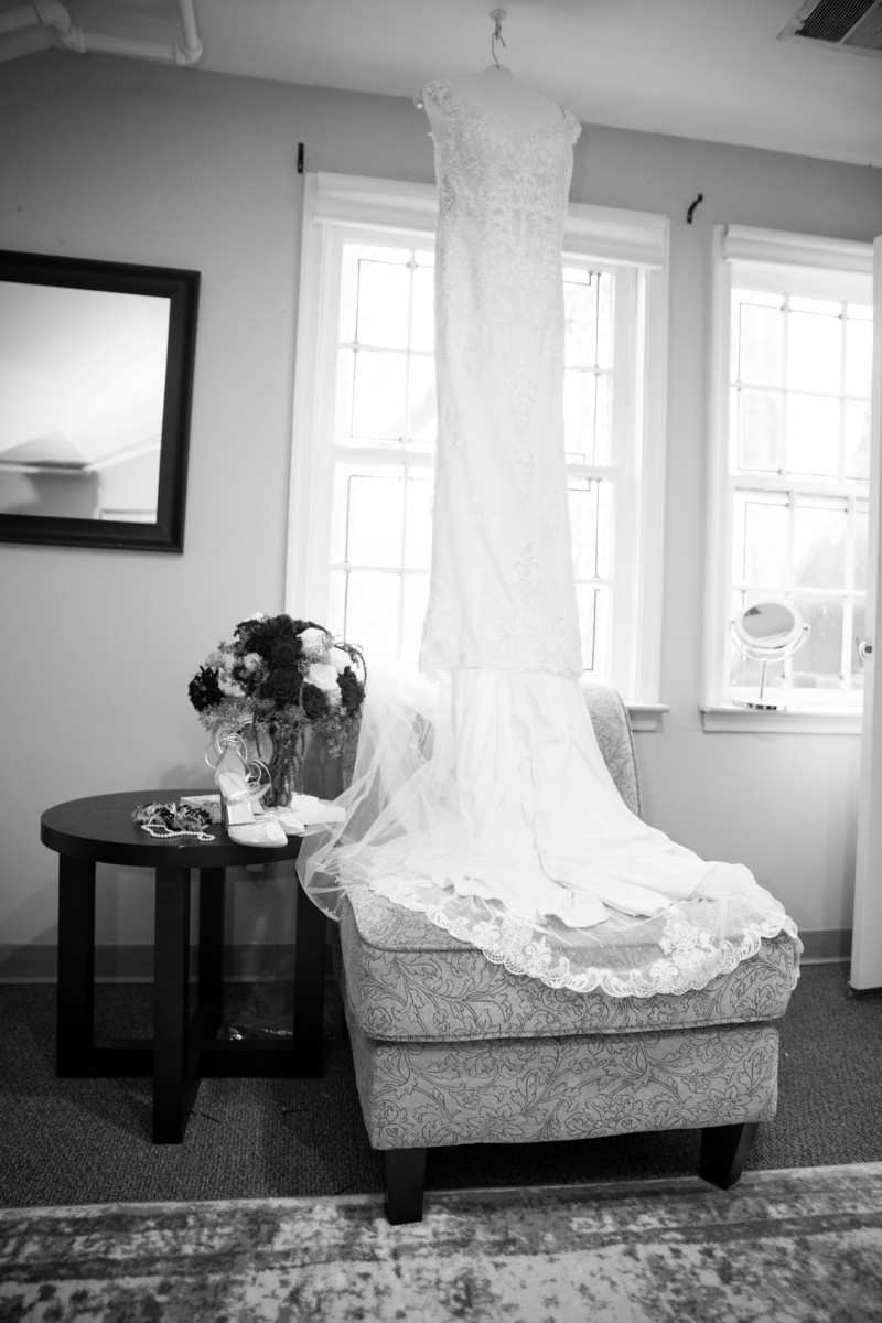 Shannon-James-Wedding-Low-Res-28 - TOMMY GATZ ENTERTAINMENT A white wedding dress hangs on a hanger in front of two windows, with a bouquet, shoes, and accessories arranged on a small table and chair nearby.