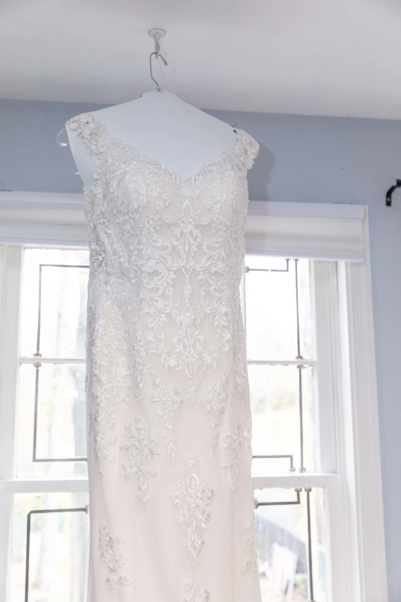 Shannon-James-Wedding-Low-Res-29 - TOMMY GATZ ENTERTAINMENT A white lace wedding dress hangs on a hanger in front of a window with light streaming in.