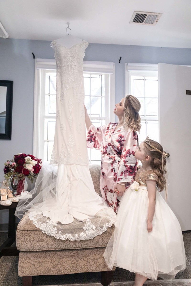 Shannon-James-Wedding-Low-Res-31 - TOMMY GATZ ENTERTAINMENT A woman and a young girl in dresses admire a hanging wedding gown in a room with large windows and floral decorations.
